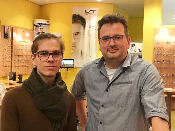 Zwei Männer mit Brille stehen in einem modernen Optiker-Geschäft mit Brillenauswahl.