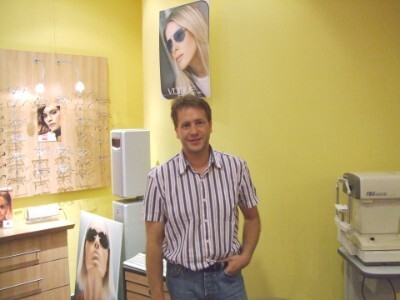 Ein freundlicher Optiker steht in einem modernen Brillengeschäft mit gelber Wand.